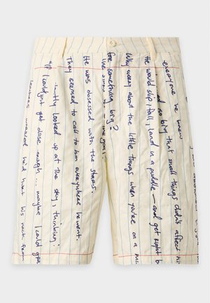 Homme portant une chemise beige à manches courtes avec un bord rouge et des illustrations d'oiseaux, un short beige avec un texte manuscrit, des chaussettes blanches et des chaussures noires.