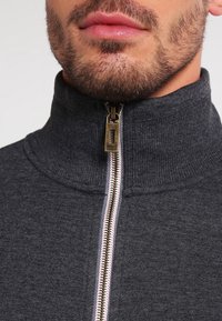 Sudadera gris con cremallera y cuello alto, con cremalleras de metal y tejido texturizado. La cremallera tiene un forro a rayas en contraste.