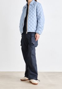Personne portant une veste matelassée bleu clair, un pantalon cargo ample bleu marine foncé avec poches, et des baskets blanches avec semelles en gomme, debout sur un sol clair.