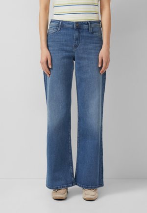 Osoba nosząca szerokie niebieskie jeansy, beżowe mokasyny oraz żółto-niebieską koszulkę w paski, stojąca na jednolitym szarym tle.