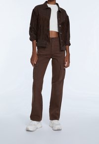 Brun denimjacka med fransdetaljer, i kombination med matchande cargobyxor och vita sneakers. Har en kort vit topp under.