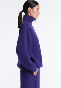 Kvinde i profil iført en lilla højhalset sweattrøje med lynlås og ribstrikkede ærmer samt matchende lilla bukser, hænderne i lommerne.