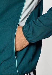 Sinihvihreä urheilutakki, jossa valkoiset yksityiskohdat; varustettu vetoketjulla suljettavalla taskulla ja kuminauhalla kiristettävillä hihoilla. Sileä pinta ja kevyt kangas.