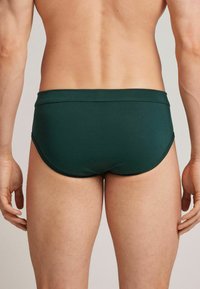 Dunkelgrüne Schwimmslips aus einem glatten, dehnbaren Stoff. Das Design zeichnet sich durch einen niedrigen Schnitt und einfache, klaren Linien am Bund aus.