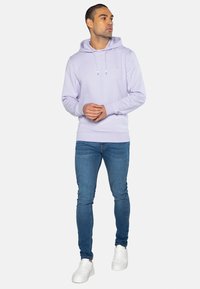 Felpa viola chiaro con cappuccio a coulisse, maniche lunghe e polsini a coste, abbinata a jeans blu aderenti e sneakers bianche.