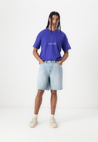T-shirt en coton violet avec le texte "HORS LIMITE", associé à un short en denim bleu clair. Le modèle porte des baskets beiges et des chaussettes blanches visibles.