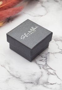 Svart texturerad smyckeskrin med fyrkantig form. Har en vit logotyp på locket. Slät yta och minimalistisk design.