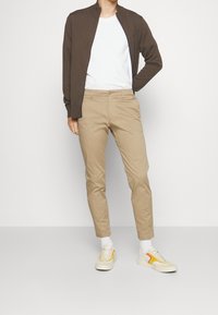 Ljust brunt bomberjacka över en vit t-shirt, matchad med beige byxor och krämfärgade sneakers med gula accenter. En enfärgad bakgrund.
