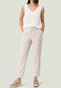 Haut sans manches blanc avec décolleté en V et bord en dentelle ; pantalons beiges ajustés avec poches ; sandales légères en brun clair avec semelles en espadrille.