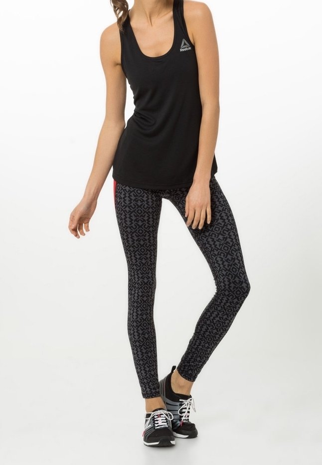 Femme portant un haut sans manches noir Reebok, des leggings noirs à motifs et des baskets noires avec des lacets blancs, debout devant un fond blanc.