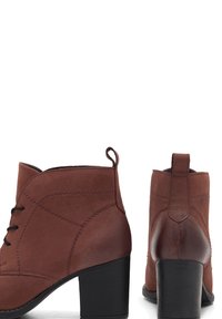 Botine din suede maro, cu o textură netedă, șireturi în față, mânere în spate și un toc solid negru. Prezintă detalii cu cusături curbate.