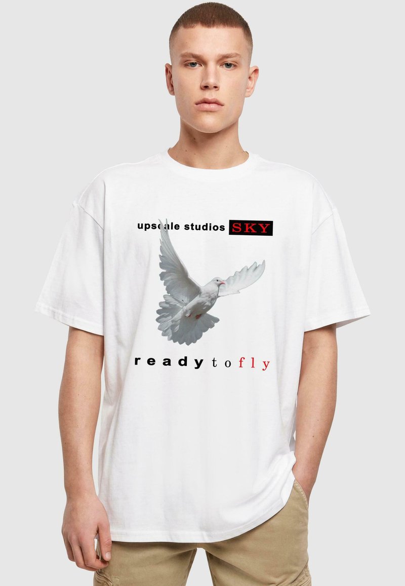 Hvid oversized t-shirt med grafik af en flyvende due og teksten "upscale studios SKY klar til at flyve" i sort og rød.