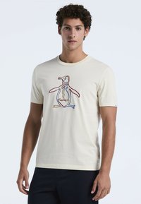 Jonge man in een crèmekleurig T-shirt met een meerkleurige geborduurde outline van een pinguïn, gecombineerd met zwarte broek, tegen een effen achtergrond.