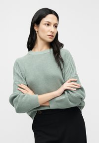 Miętowo zielony sweter z dzianiny z okrągłym dekoltem i prążkowanymi mankietami. Tekstura jest miękka i elastyczna, zestawiona z czarnymi spodniami z wysokim stanem.
