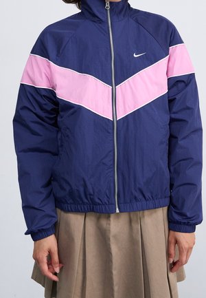 Osoba w granatowej kurtce na zamek błyskawiczny z różowym paskiem w kształcie litery V i beżową plisowaną spódnicą, ręce swobodnie opuszczone wzdłuż boków.