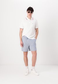 Hvit polo skjorte med røde detaljer, lyseblå stripete shorts og hvite sneakers. Modellen bærer solbriller og står mot en enkel bakgrunn.