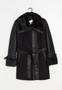 Abrigo de invierno negro con mangas de cuero acolchado, cuello y puños de piel, cintura ceñida con cinturón y cierre frontal con cremallera colgado de una percha negra.