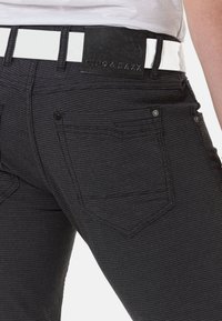 Schwarze kariert Denim-Jeans mit einem weißen strukturierten Gürtel. Merkmale sind Gesäßtaschen und ein geprägtes Lederpatch mit Markenlogo.