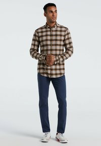 Man staat met gevouwen handen, draagt een bruin geruit overhemd met knopen, marineblauwe broek en witte sneakers voor een effen achtergrond.