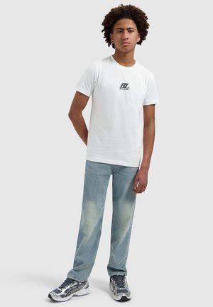 Jonge man met krullend haar die een wit T-shirt met Ballin-logo draagt, lichtblauwe spijkerbroek en grijs-witte sneakers, staand met één hand achter zijn rug.