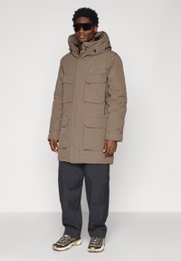 Brun, isoleret parka med hætte og flere lommer foran, kombineret med sorte cargobukser og teksturerede sneakers med grå accenter.