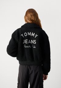 Veste en fausse fourrure noire avec un design court, des manches oversize et un ourlet côtelé. Le dos arbore l'inscription "TOMMY JEANS" en broderie blanche.