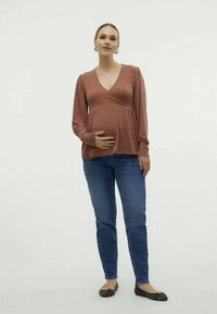 T-shirt de maternidade em rust com decote em V cruzado, mangas longas; combinado com jeans azuis e sapatos planos em padrão preto e branco. Tecido simples e suave.