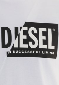 Camiseta de algodón blanca con un logo negro de "DIESEL" y la frase "PARA UNA VIDA EXITOSA". Presenta una costura diagonal. Textura suave.