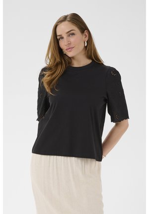 Hosszú világosbarna hajú nő fekete csipkés rövid ujjú felsőt és világos bézs szoknyát visel, sima háttér előtt áll.