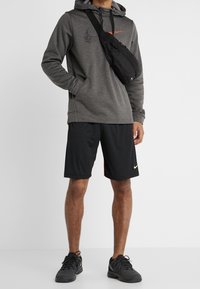 Sweatshirt à capuche gris avec des accents noirs, associé à des shorts de sport noirs avec des détails rouges. Sac bandoulière noir et chaussures de sport.