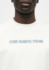 Κοντινό πλάνο ενός ατόμου που φοράει λευκό μπλουζάκι με το κείμενο "THE NORTH FACE" σε στυλιζαρισμένα φωτεινά γαλάζια γράμματα σε σχήμα φυσαλίδων στο στήθος.