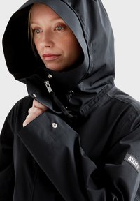 Veste étanche noire avec capuche, dotée d'une fermeture éclair, de boutons pression et d'un patch logo réfléchissant. Texture lisse et design ajusté.