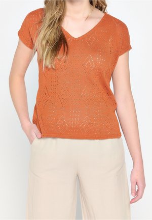 Kvinde iført kortærmet orange strikbluse med geometriske mønstre og lyse beige bukser, hånd i lommen, foran ensfarvet baggrund.