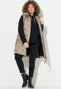 Beige Pufferweste mit abstrakten Linienausschnitten, Kapuze, Reißverschluss vorne und schwarzen Akzenten. Tragen über einem schwarzen Oberteil und schwarzen Jeans mit Stiefeletten.