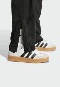 Baskets blanches avec trois rayures noires, une semelle en gomme et un capuchon en daim. Pantalons de survêtement noirs avec des zips sur les côtés et des accents côtelés.