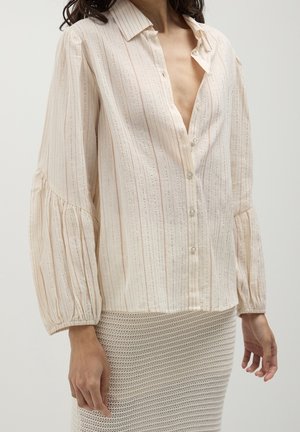 Femme portant un chemisier beige clair à rayures, boutonné, avec de longues manches bouffantes, et une jupe beige texturée.