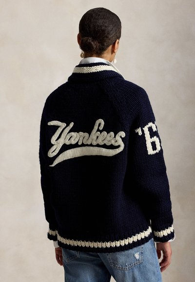 Personne avec les cheveux en chignon portant une veste en maille bleu marine avec le lettrage blanc « Yankees » et le numéro « 6 » sur la manche, associée à un jean bleu.