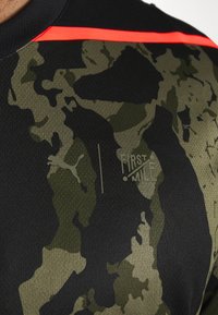 Primer plano de una camisa de camuflaje negra y verde con una franja roja, que presenta los logotipos de Puma y First Mile en el pecho.