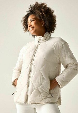 Donna sorridente che indossa una giacca trapuntata color crema con collo alto, mani nelle tasche con zip e pantaloni bianchi su sfondo uniforme.