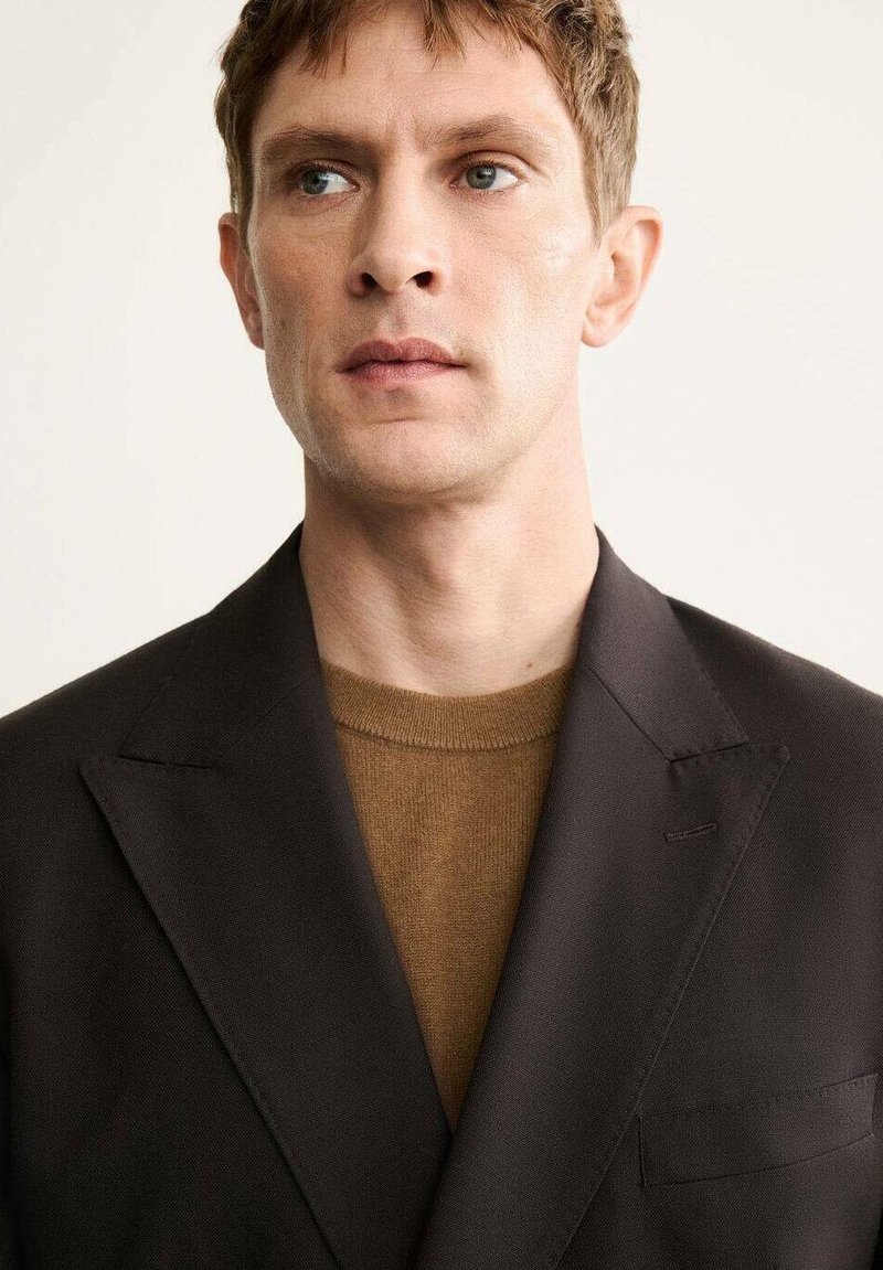 Black suit jacket with notch lapels over a brown sweater. Smooth fabric with structured fit, single chest pocket, and minimal details.