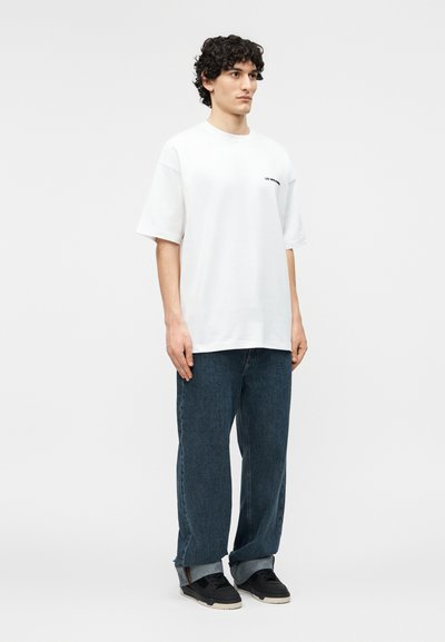 Άνδρας με σγουρά μαλλιά, που φοράει χαλαρό λευκό μπλουζάκι, φαρδιά μπλε τζιν με ρεβέρ στο κάτω μέρος και μαύρα αθλητικά, στέκεται μπροστά σε λευκό φόντο.