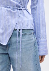 Φωτεινό γαλάζιο πουκάμισο τύπου wrap με ρίγες και ζώνη στη μέση, συνδυασμένο με γαλάζιο τζιν. Μακριά μανίκια, λεία υφή υφάσματος και εφαρμοστή γραμμή.