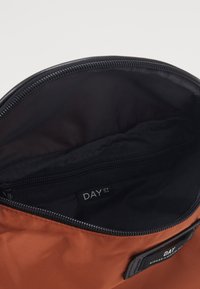 Brązowa torba tekstylna z czarnym zamkiem błyskawicznym, wyposażona w nylonowe wnętrze oraz etykietę z napisem "DAY ET". Gładka tekstura, zaokrąglony kształt.