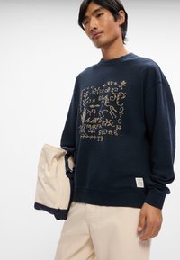 Σκούφος ναυτικού σε μαλακό ύφασμα με υφή χρυσού γραφικού σχεδίου. Διαθέτει ριμπ μανσέτες και στρίφωμα, με μια μικρή λεπτομέρεια ετικέτας στο κάτω μέρος.