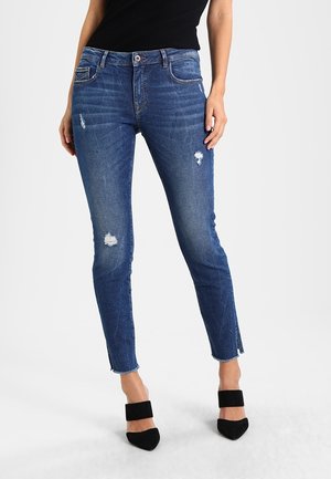 Jeans de mezclilla azules ajustados con detalles desgastados, dobladillos deshilachados, cinco bolsillos y cierre de botón. Llevados con tacones negros con tira en el tobillo.