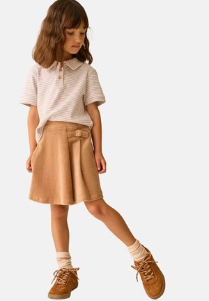 Nuori tyttö seisoo yhdellä jalalla ojennettuna, päällään beige ruudullinen paita, vaaleanruskea corduroyperhonen vyöllä, vaaleancremekorkeat sukat ja ruskeat kengät.