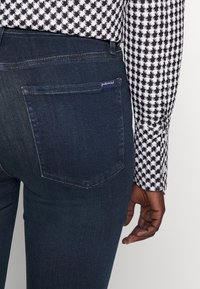 Pantalones de mezclilla de color azul oscuro con un bolsillo trasero y una camisa de manga larga con patrón de espiga en blanco y negro, con la mano descansando a un lado.