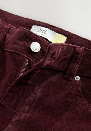 Burgundské džíny s mírným vzestupem, slim fit, s kovovým zapínáním, viditelným zipem a měkkou texturou látky. Etiketa uvádí "Slim Mid Rise, Vyrobeno v Kambodži."