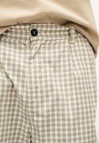 Μπεζ και λευκό καρό παντελόνι με μαύρο κουμπί και θηλιές για ζώνη, φορεμένο με μπεζ τοπ που είναι μέσα στο παντελόνι.