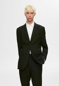 Ciemnoszary żakiet i pasujące spodnie, z klapami, dwoma guzikami i kieszeniami z przodu. Biała koszula pod spodem dopełnia całość.